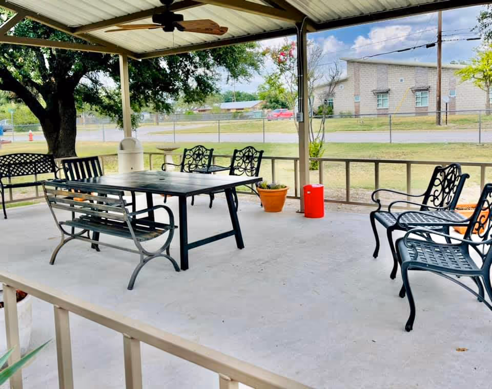 Covered outdoor patio area with metal benches and chairs arranged around a rectangular table. There are potted plants and a ceiling fan under the roof. In the background, there is a large tree, a fence, and a building across the street under a partly cloudy sky.