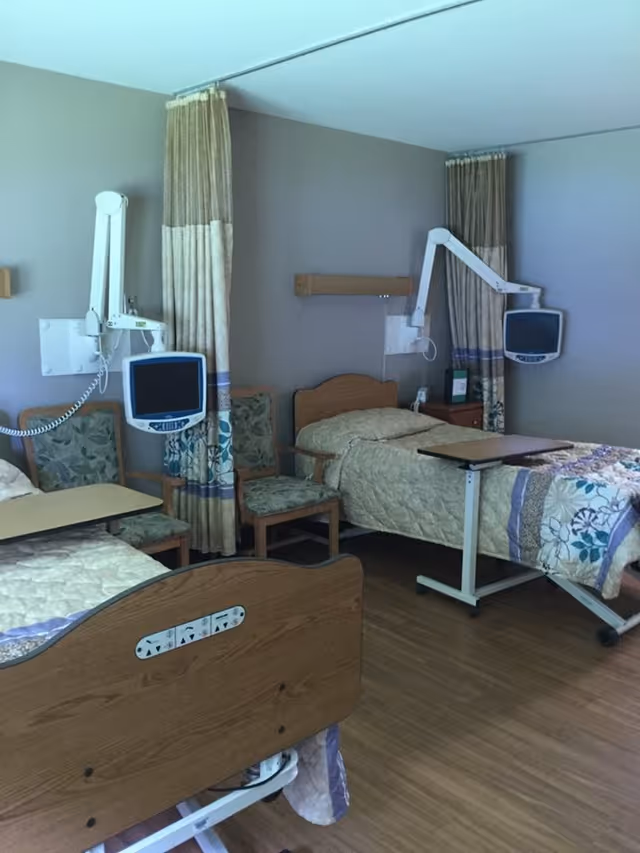 A healthcare facility room with two hospital beds, each equipped with a mounted monitor and an overbed table. There are two chairs with floral upholstery between the beds, and privacy curtains hanging on the wall behind the beds. The room has wood flooring and neutral-colored walls.