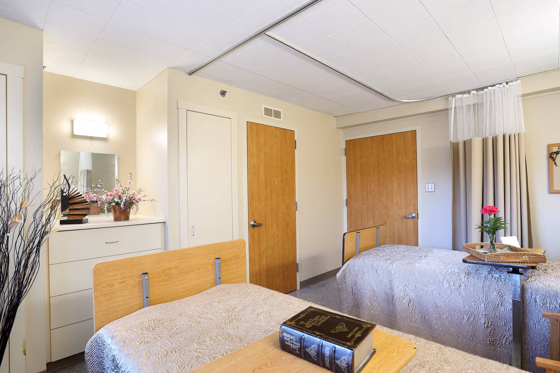 Two-bed senior living bedroom with wooden headboards, a dresser topped with flowers, and a tray on one bed.