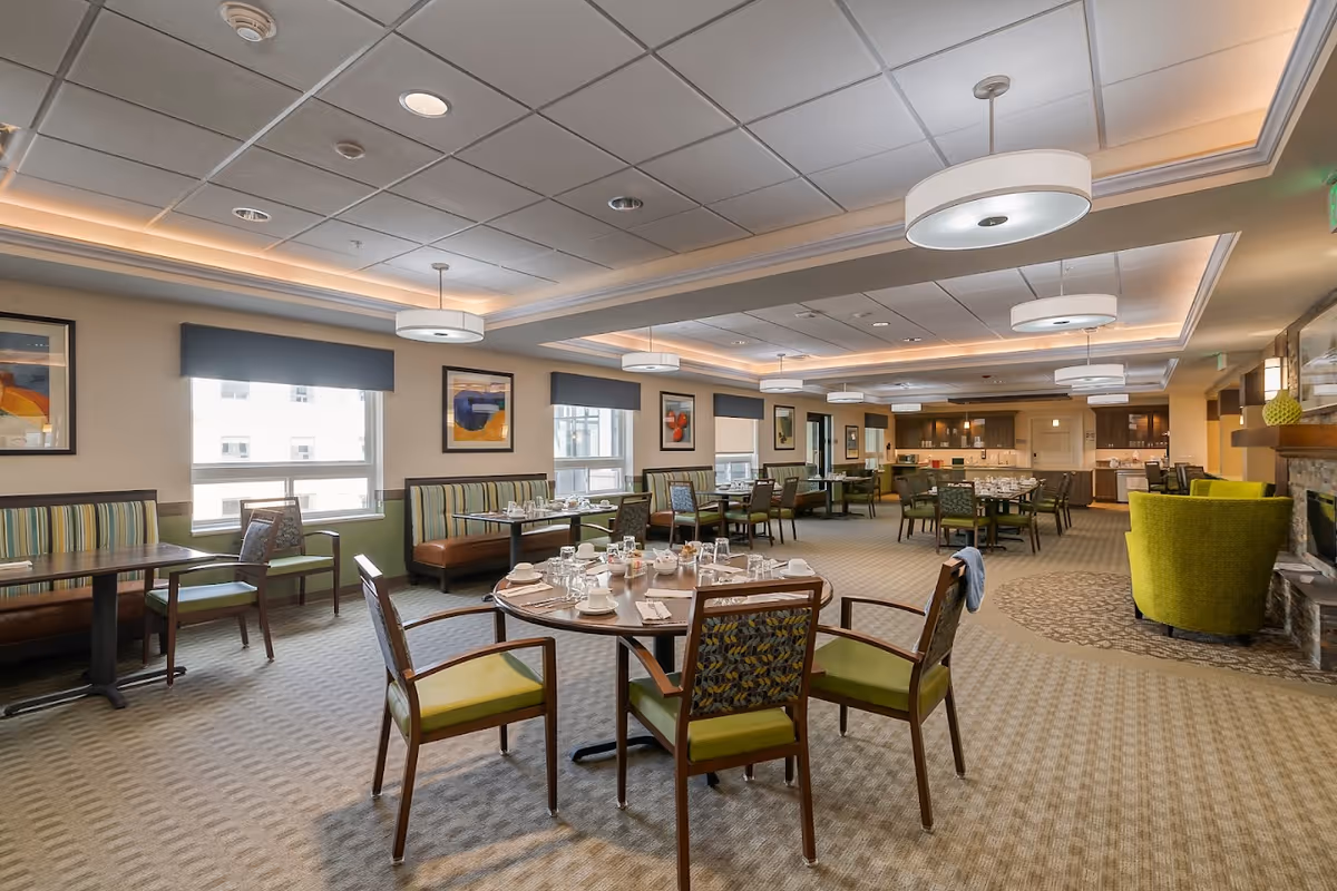 A spacious dining room in a senior living facility with multiple tables and chairs arranged neatly. The tables are set with cups, glasses, and napkins. The room features large windows with blue valances, framed artwork on the walls, and modern ceiling lights. There is a seating area with green armchairs near a stone fireplace on the right side of the room.