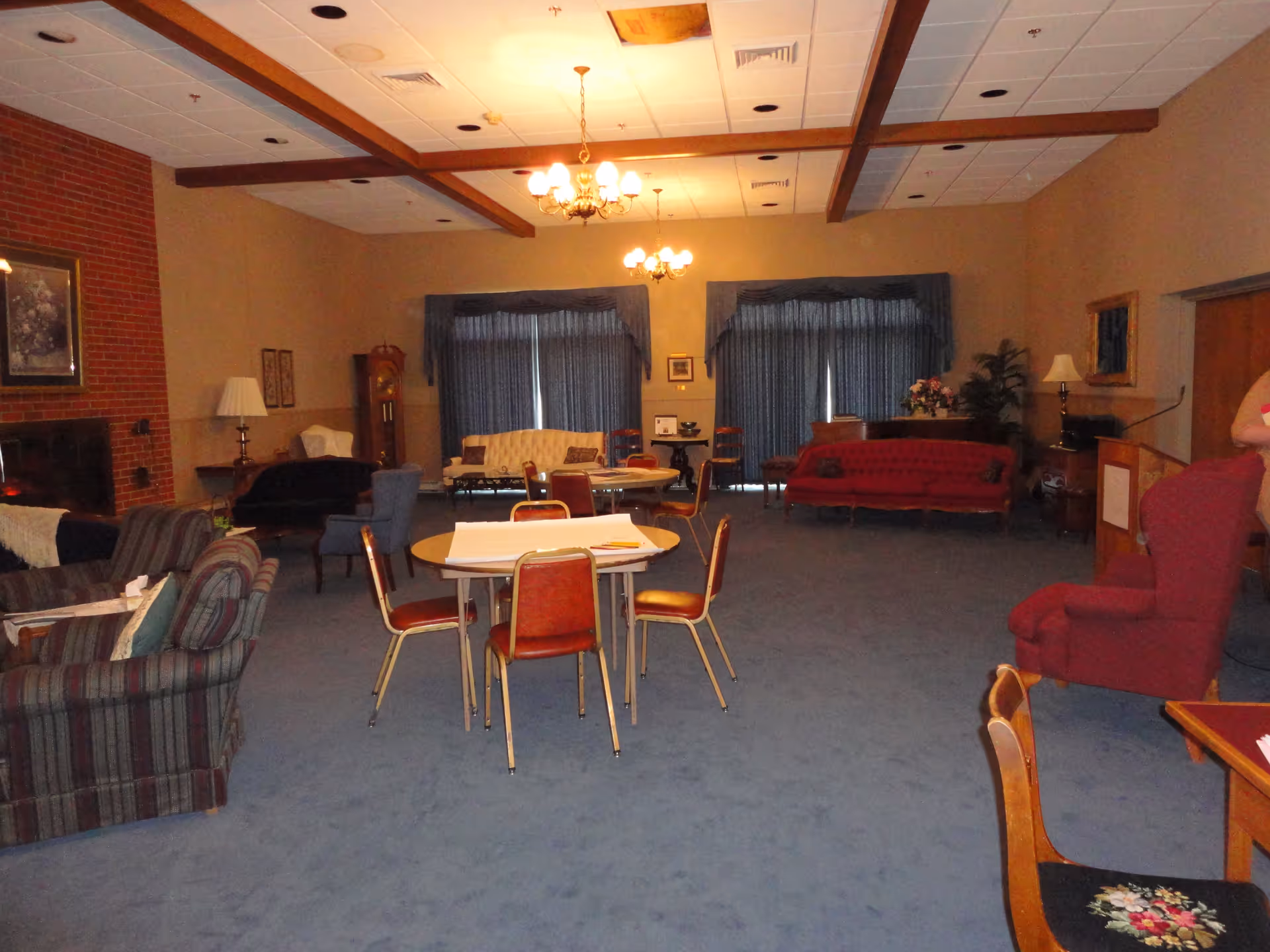 A spacious living room with blue carpet, multiple seating areas including sofas, armchairs, and small round tables with chairs. The room features a brick fireplace on the left wall, two chandeliers hanging from the ceiling, and large windows covered with blue curtains at the back. There are various lamps, framed pictures on the walls, and a grandfather clock near the back left corner.