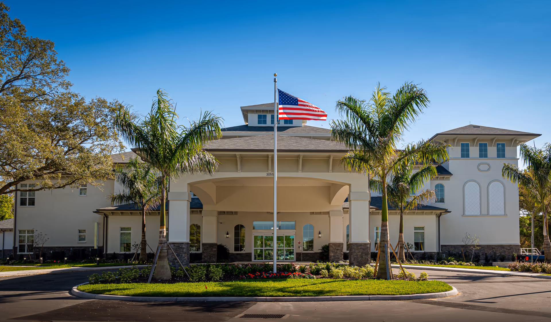 Front exterior view of a large, modern senior living facility building with a covered entrance, palm trees, an American flag on a flagpole, and clear blue sky.