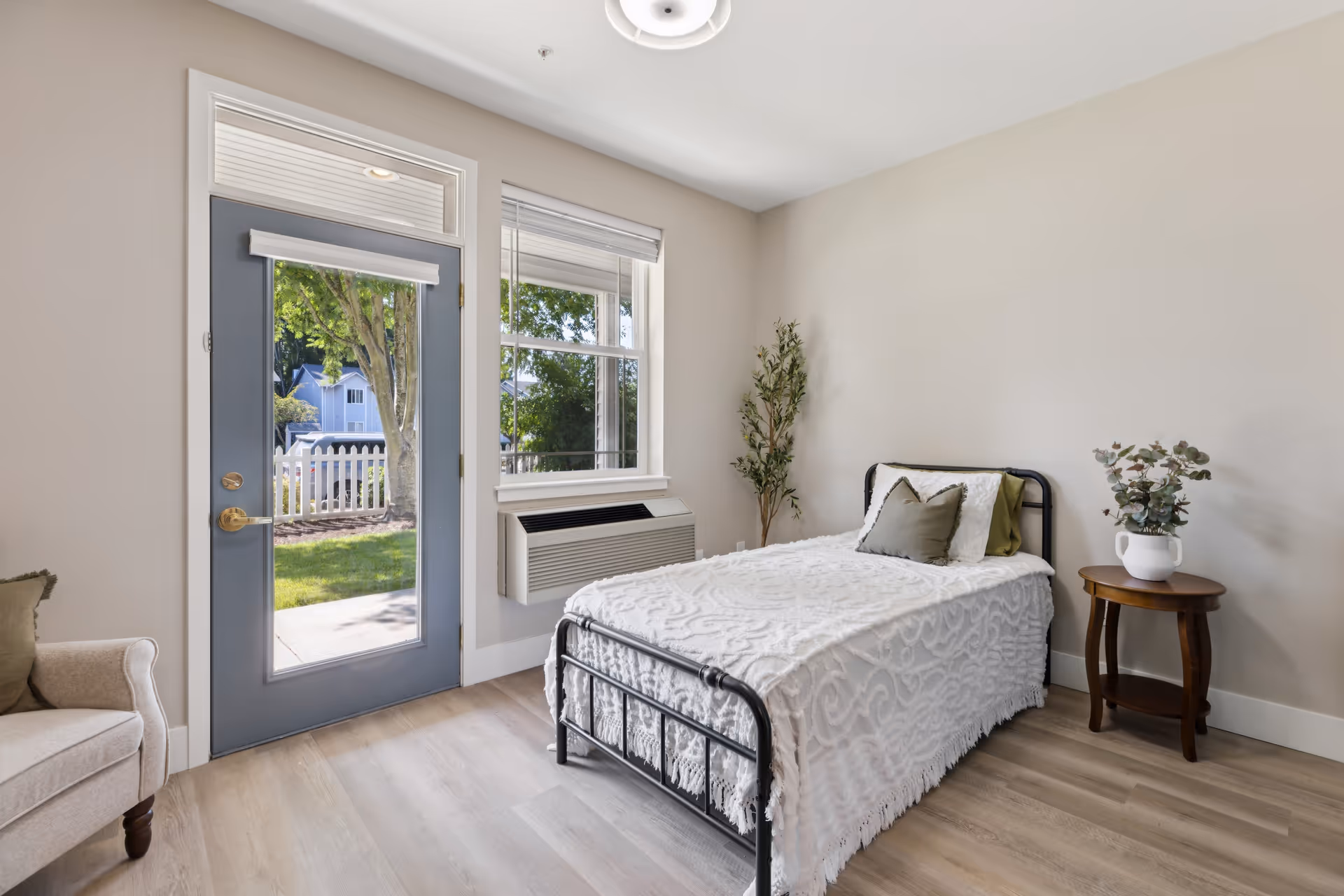 A bright and cozy bedroom with a single metal bed covered in a white textured blanket and two pillows. Next to the bed is a small wooden round side table with a white vase holding green foliage. There is a beige armchair partially visible on the left side. The room has light wood flooring, beige walls, a large window, and a glass door leading outside to a green yard with trees and a white picket fence.