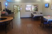 Interior view of a nursing home common area with wooden flooring, tables and chairs arranged for seating, a coffee station on the left, a hallway leading to other rooms, and a wall-mounted television on the right.