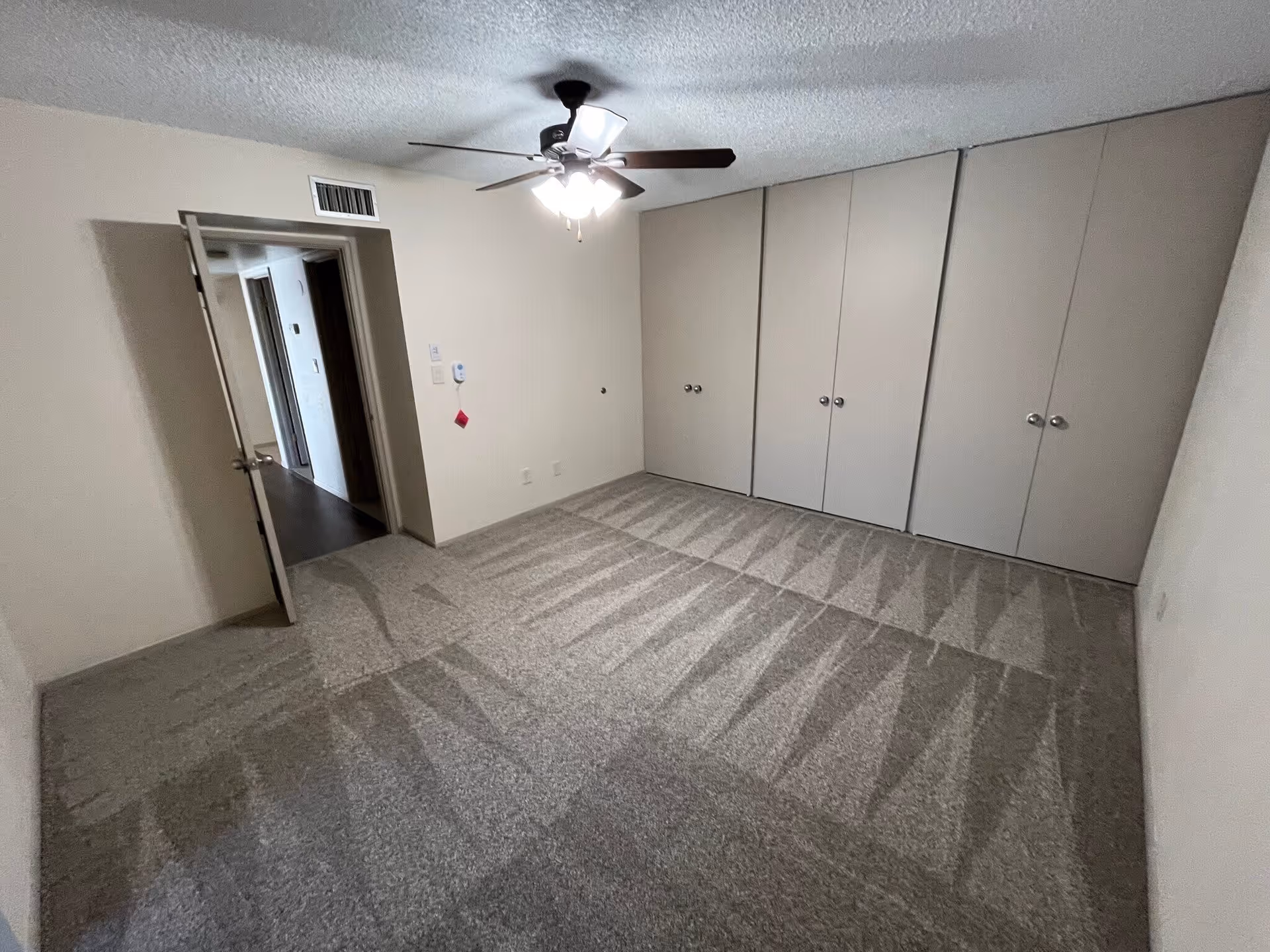 Empty bedroom with beige carpet, a ceiling fan with lights, a row of closed closet doors, and an open door leading to a hallway.