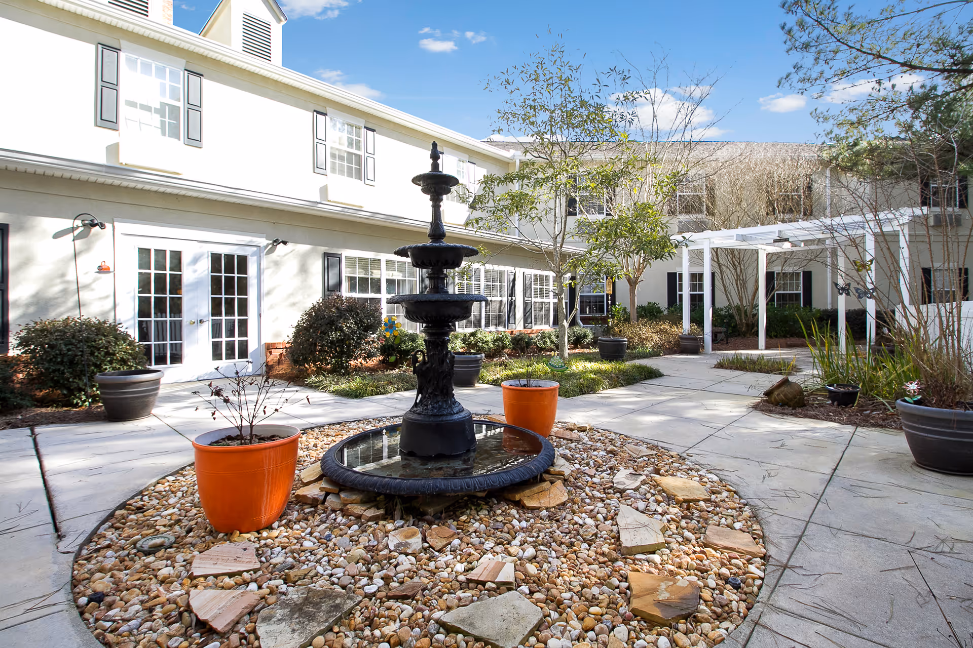 Outdoor courtyard area of a senior living facility with a black tiered water fountain in the center surrounded by rocks and two orange plant pots. The courtyard is paved with concrete walkways and has various plants and small trees. The building surrounding the courtyard is two stories with white walls and multiple windows and doors. The sky is clear and blue.