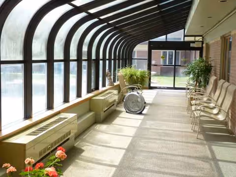 Sunlit enclosed hallway with chairs, plants, and a wheelchair near large glass windows.