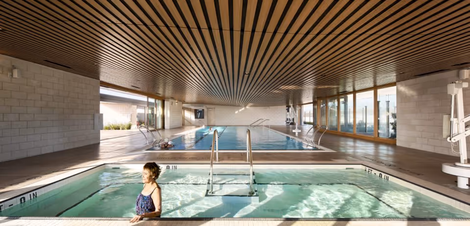 Indoor swimming pool area with a smaller hot tub in the foreground where a woman is standing in the water. The space features a wooden slatted ceiling, large windows on the right side, and light-colored brick walls. The main pool is in the background with metal handrails and pool equipment visible on the sides.