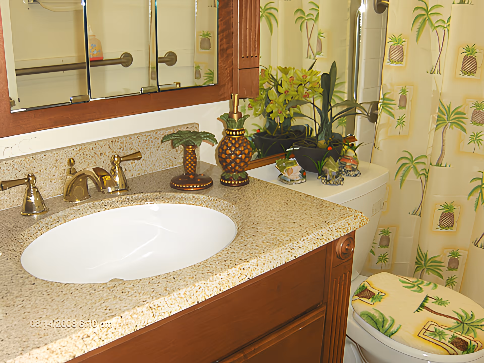 Bathroom vanity with an oval sink, brass faucet, decorative plants, and a toilet beside a palm-patterned shower curtain.