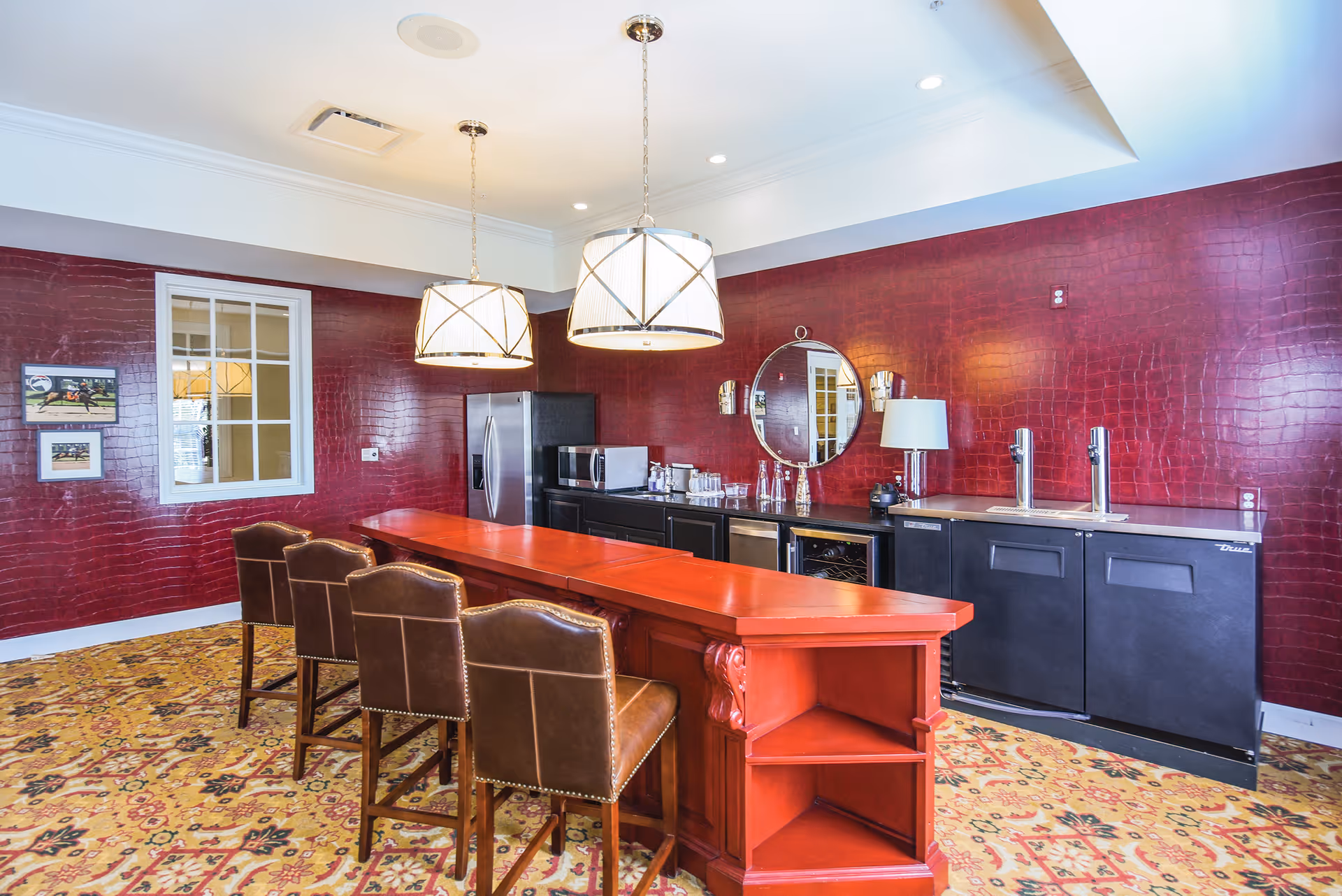 Interior view of a senior living facility's kitchen area with a long red wooden island counter surrounded by four brown leather chairs. The back wall is covered in a textured dark red wallpaper, featuring a round mirror, two wall sconces, a lamp, and kitchen appliances including a refrigerator, microwave, and beverage dispensers. Two large pendant lights hang from the ceiling, and the floor is covered with a patterned carpet.