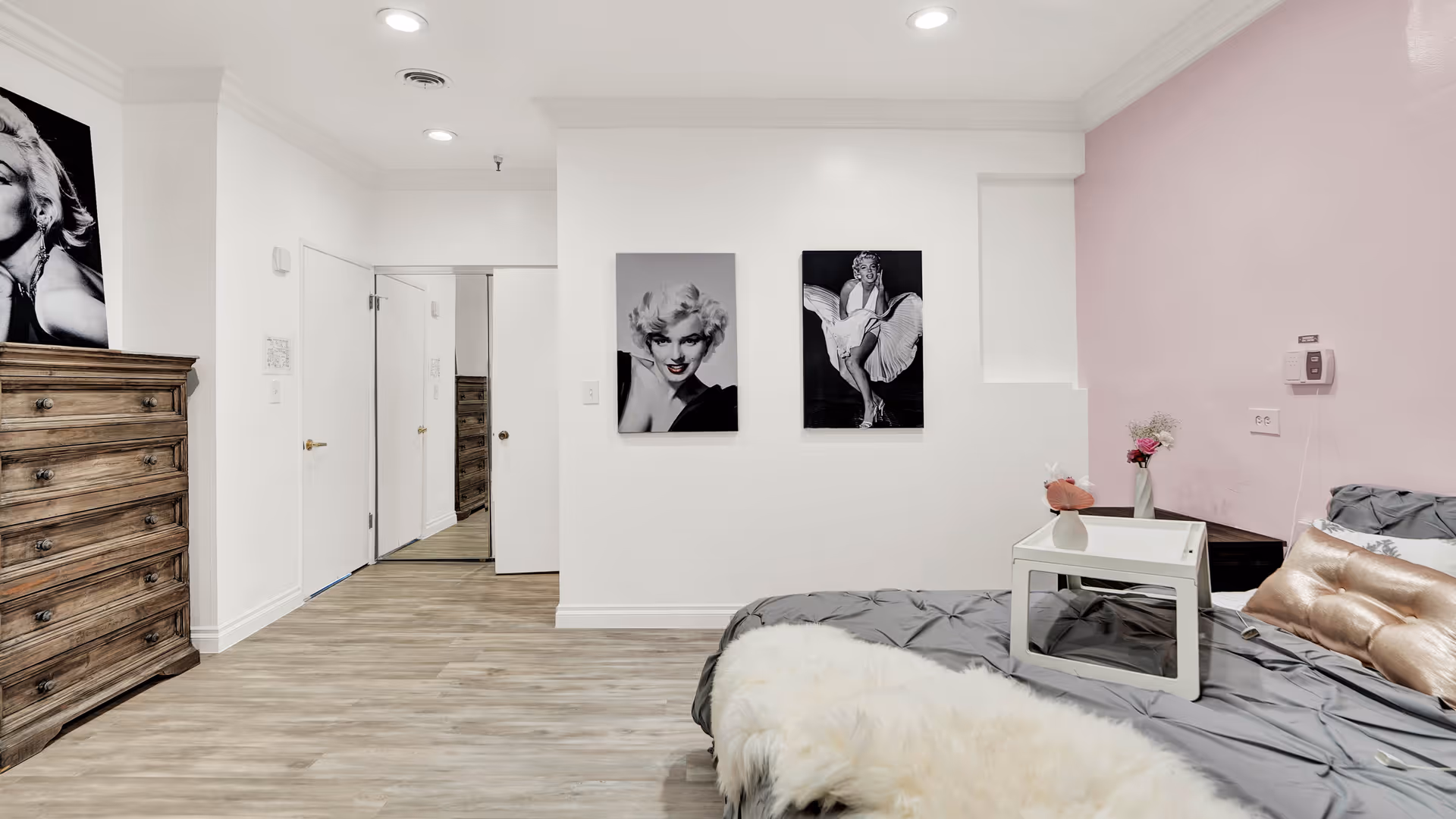 A bedroom with light wood flooring, white walls, and one pink accent wall. The room features a bed with gray bedding, a white tray table, a gold pillow, and a white fluffy throw. There is a wooden dresser on the left side and two black and white portraits of Marilyn Monroe on the wall. A door with a mirror is visible in the background.