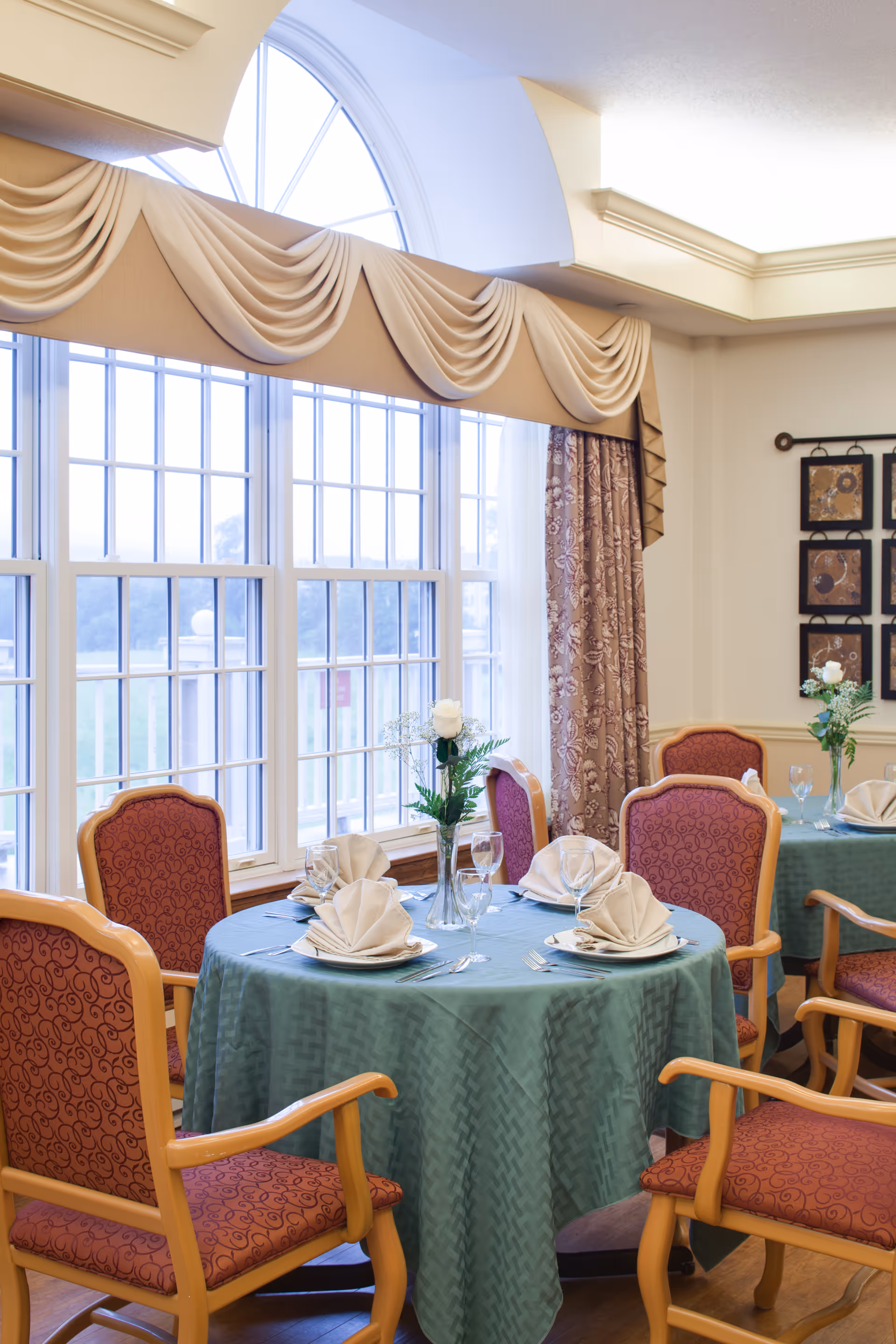 A dining room with round tables covered in green tablecloths, each set with folded napkins, plates, silverware, and wine glasses. The chairs have wooden frames with red patterned upholstery. Large windows with decorative drapes allow natural light to fill the room. A vase with a white rose and greenery is placed in the center of each table.