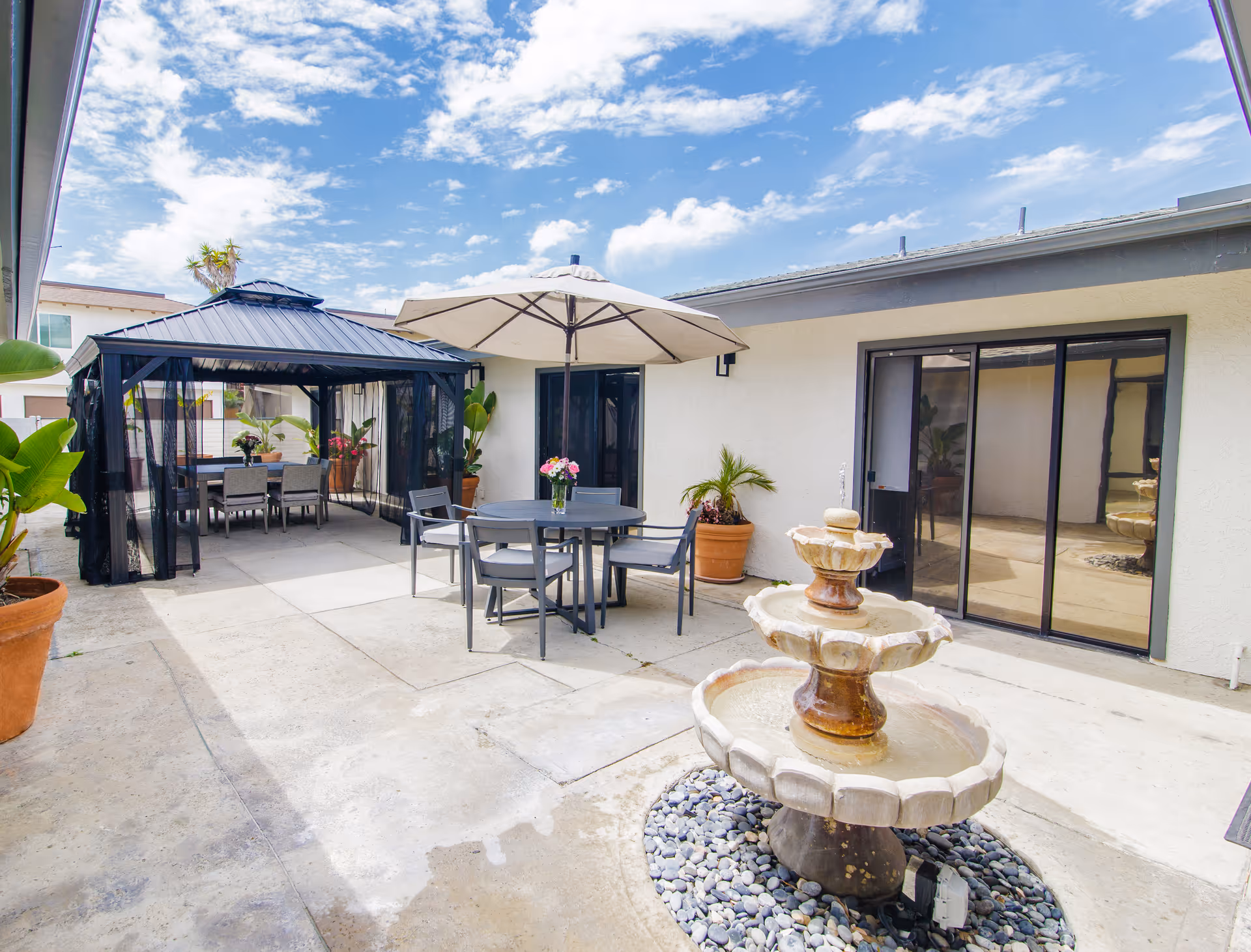 Outdoor patio area with a three-tier stone water fountain surrounded by pebbles, a round table with four chairs under a large beige umbrella, and a black gazebo with a dining table and chairs inside. Potted plants are placed around the patio, and sliding glass doors lead into the building. The sky is partly cloudy.