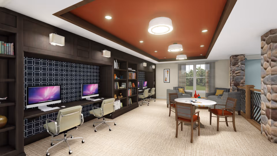 A modern communal room featuring a row of computer workstations with swivel chairs along a dark wood built-in shelving unit. The room has a beige carpet, a round table with four chairs in the center, and a seating area with a sofa and armchairs near a window. The ceiling has recessed lighting and a reddish-brown accent panel with circular light fixtures. Stone pillars and a railing are visible on the right side.