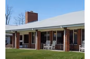Single-story brick building with a covered porch featuring white rocking chairs, a chimney, and a green lawn in front under a clear blue sky.