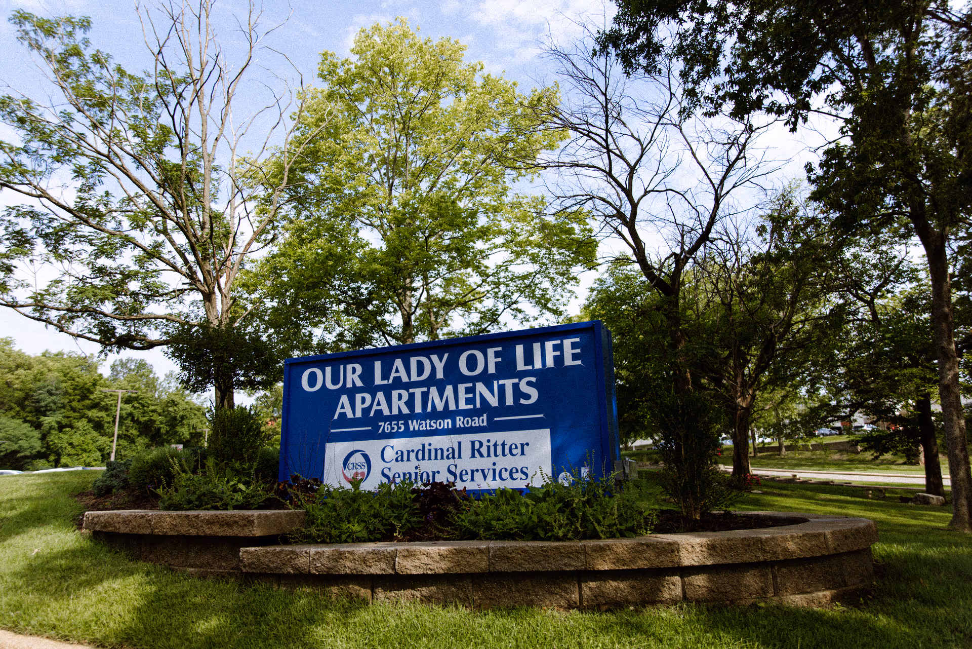 Blue entrance sign reading 'Our Lady of Life Apartments' set in landscaped greenery and trees.