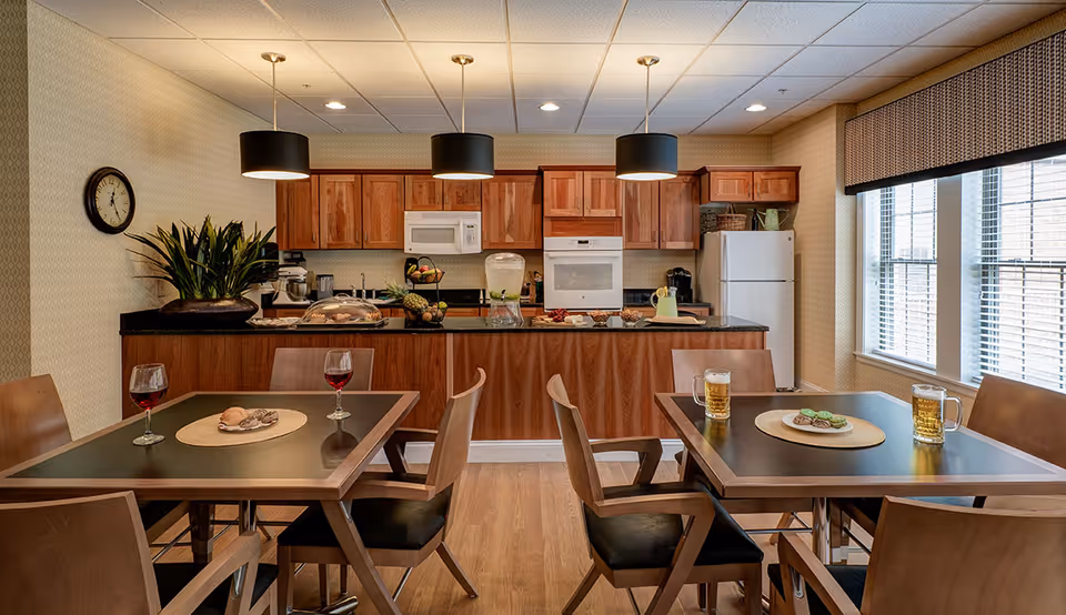 A cozy kitchen and dining area in a senior living facility featuring wooden cabinets, a white refrigerator, microwave, and oven. There are two square dining tables with wooden chairs, each table set with drinks and small plates of food. The room is well-lit with three pendant lights hanging above the kitchen counter and natural light coming through large windows with blinds.