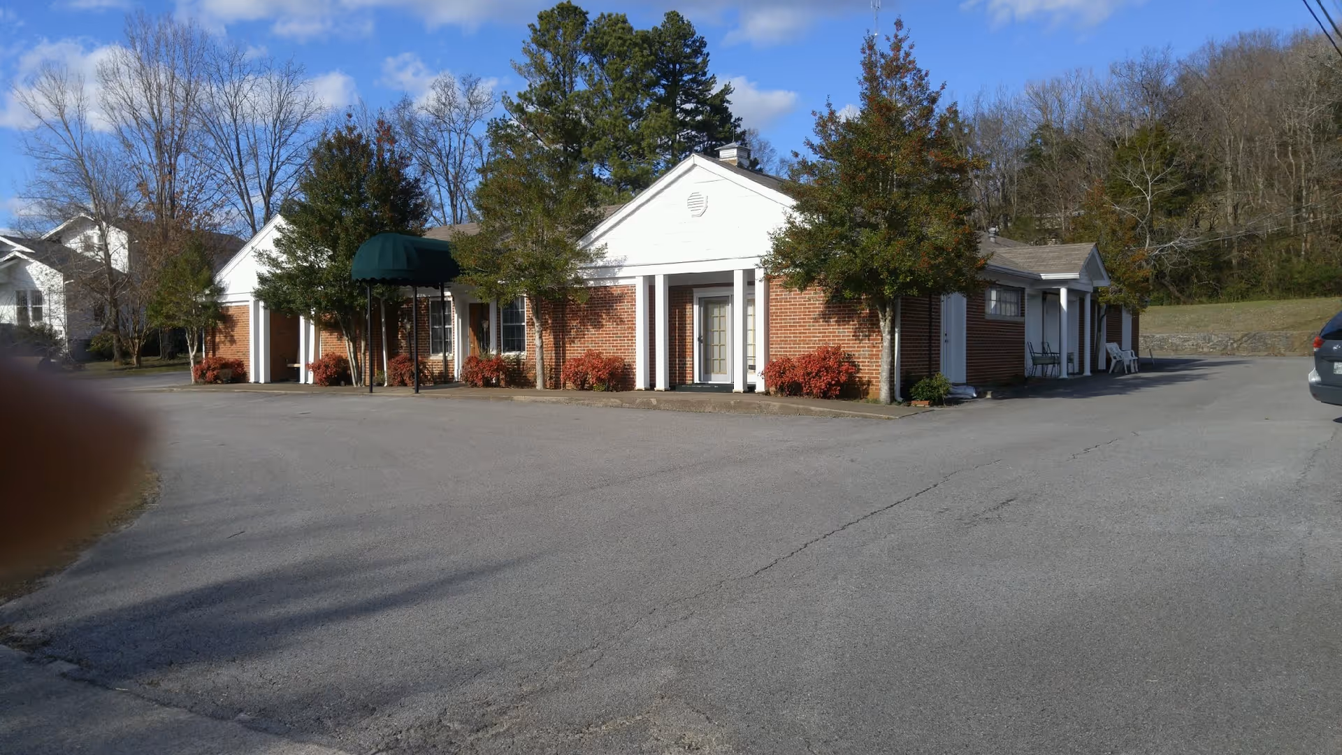 Exterior view of a single-story brick building with white trim and columns, surrounded by trees and shrubs. The building has a green awning over one entrance and a paved driveway in front. There is a car parked on the right side and a finger partially visible on the left side of the image.