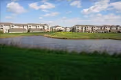 Grassy foreground and a pond with apartment-style community buildings under a partly cloudy blue sky.