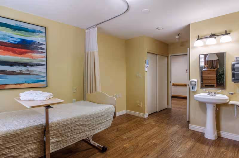 A rehabilitation or nursing facility room with a hospital bed covered in a beige blanket, a small overbed table with folded towels, a wall-mounted sink with a mirror and soap dispenser, a privacy curtain on a ceiling track, wooden flooring, and a colorful abstract painting on the wall.