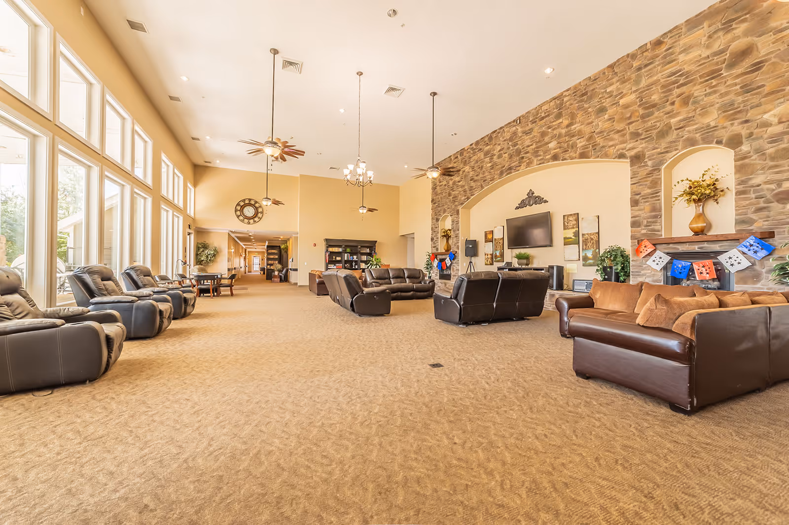 Spacious senior living facility common area with large windows on the left letting in natural light. The room features multiple leather recliners and sofas arranged around a stone accent wall with a mounted flat-screen TV and decorative artwork. Ceiling fans and pendant lights hang from the high ceiling, and there is a large clock on the far wall above a hallway entrance.