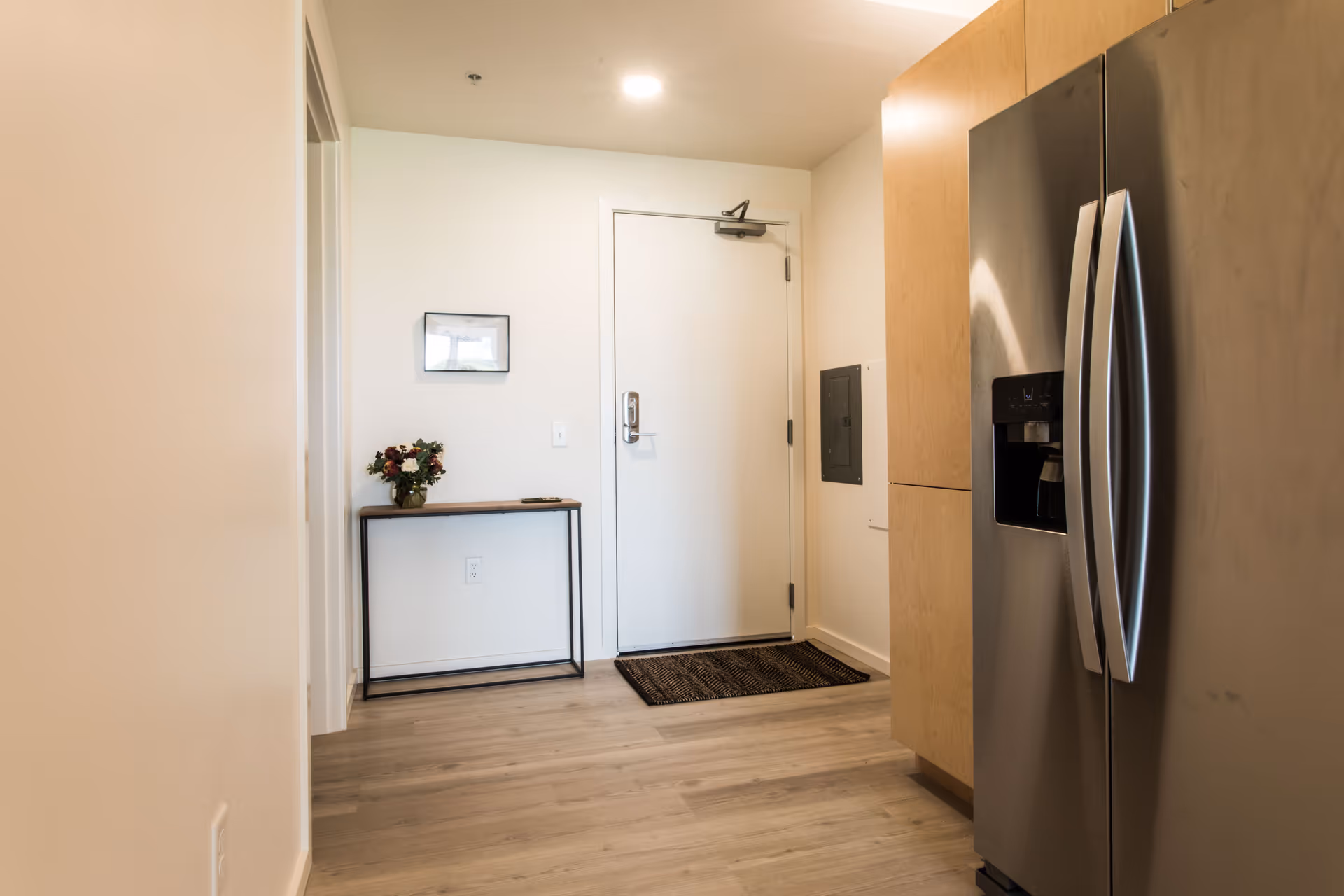 Apartment entryway with a white front door, a small console table with flowers, and a stainless steel refrigerator visible on the right.