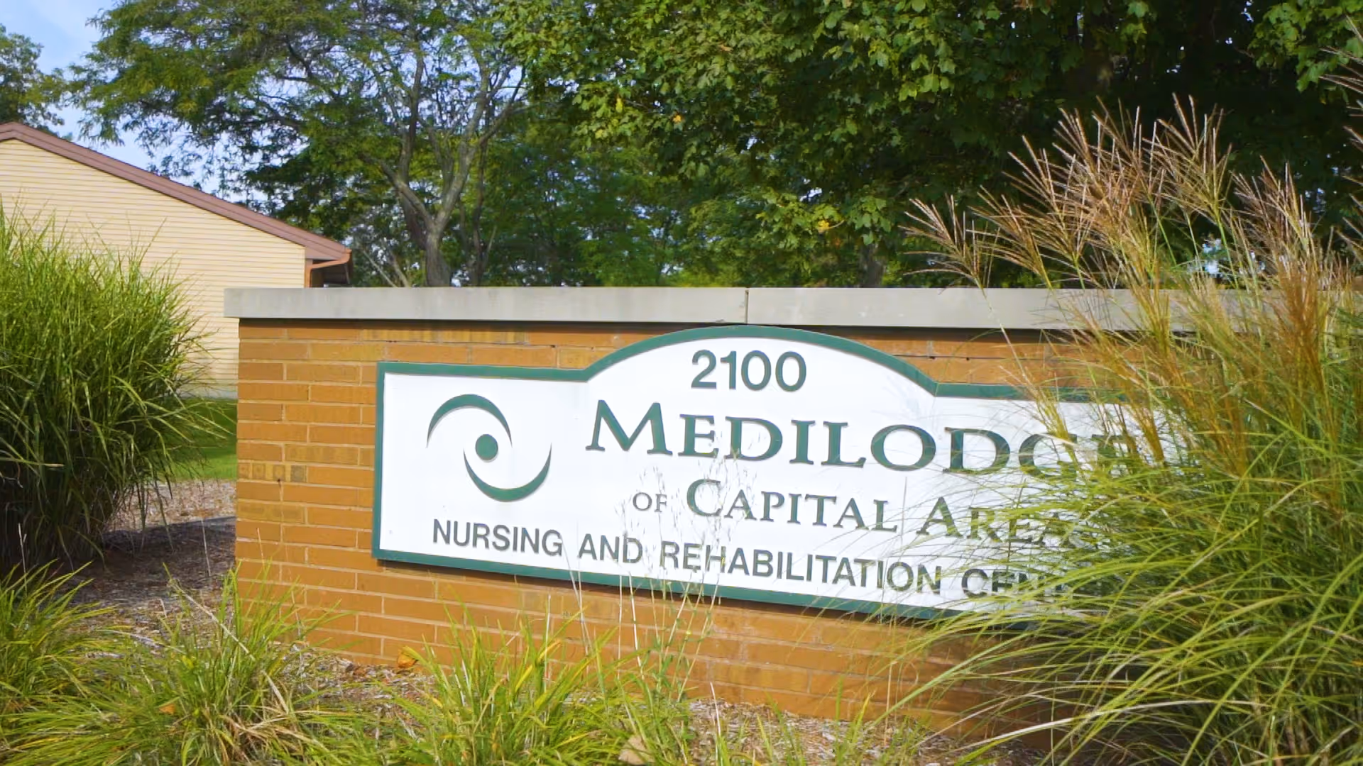 Brick entrance sign reading 'Medilodge of Capital Area' with the address 2100, surrounded by ornamental grasses and trees.