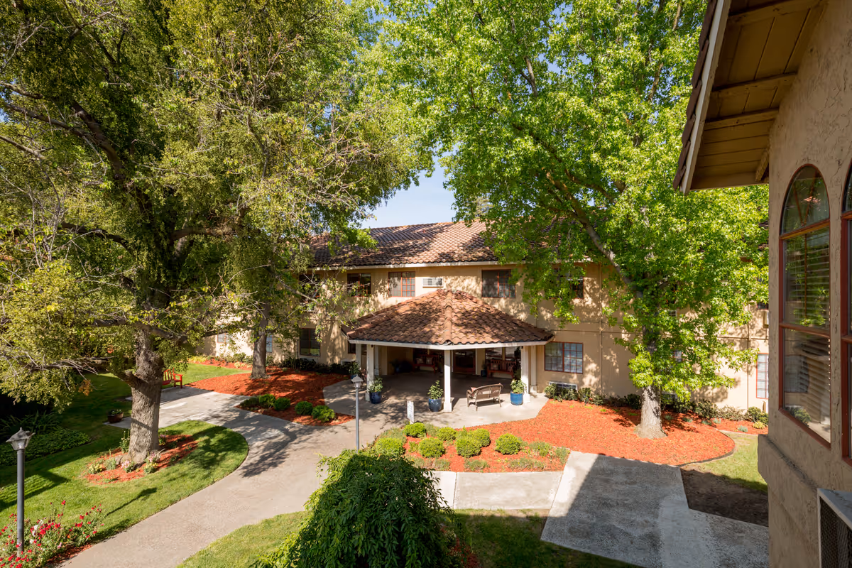 Front entrance of a two-story residential building with a tiled canopy, large trees, a circular driveway, and landscaped grounds.