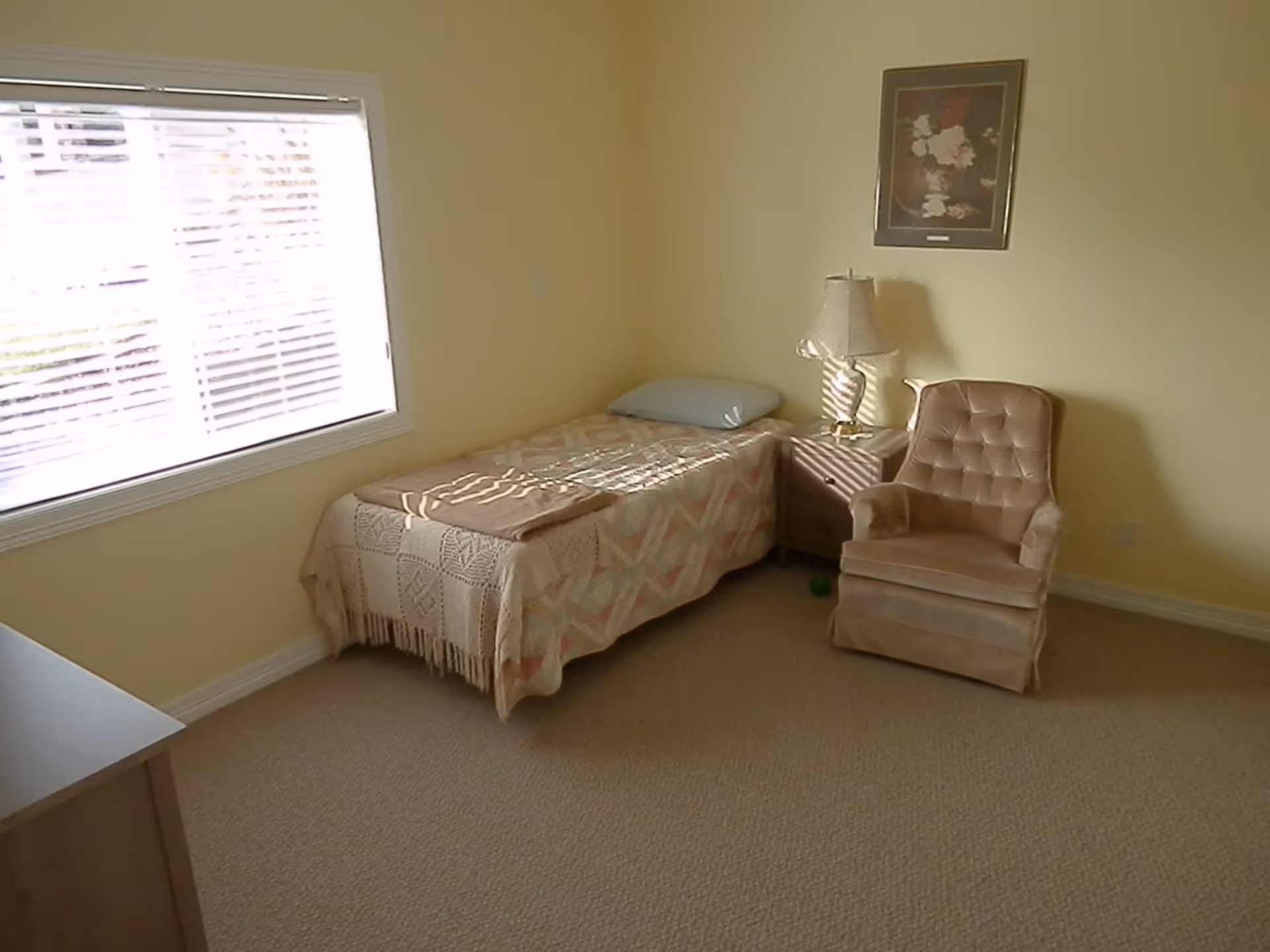 A simple bedroom with a single bed covered with a patterned blanket and a pillow, a beige armchair, a small nightstand with a lamp, and a framed floral picture on the wall. There is a window with blinds letting in natural light.