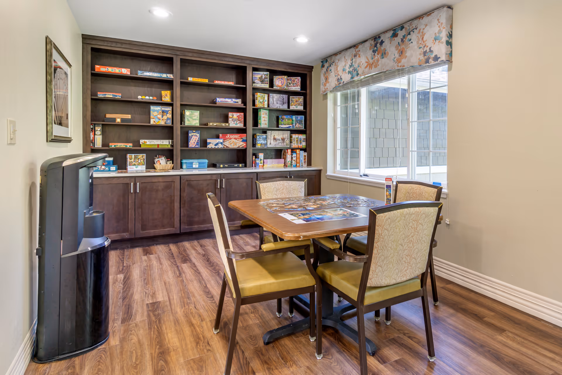 A well-lit room with a wooden table and four chairs arranged around it. On the table, there is a partially completed jigsaw puzzle. Behind the table, there is a large dark wooden cabinet with shelves filled with various board games and puzzles. A large window with floral valance lets in natural light, and the floor is covered with wood-patterned flooring. A black water dispenser is positioned against the left wall.