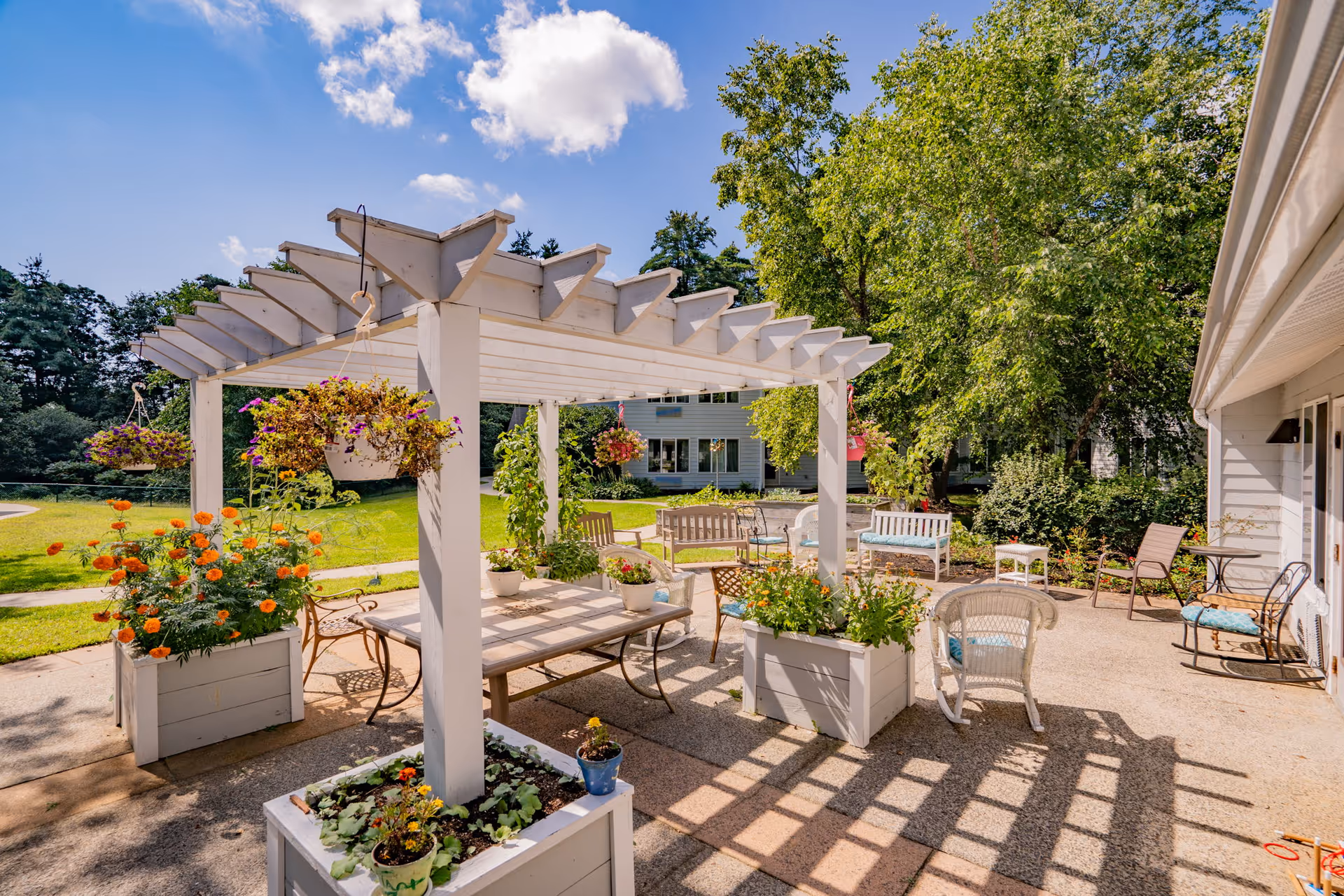 Outdoor patio area at Birchwoods at Canco Senior Living featuring a white pergola with hanging flower pots, various potted plants, multiple seating options including benches, chairs, and a table, surrounded by green trees and a lawn under a blue sky with some clouds.