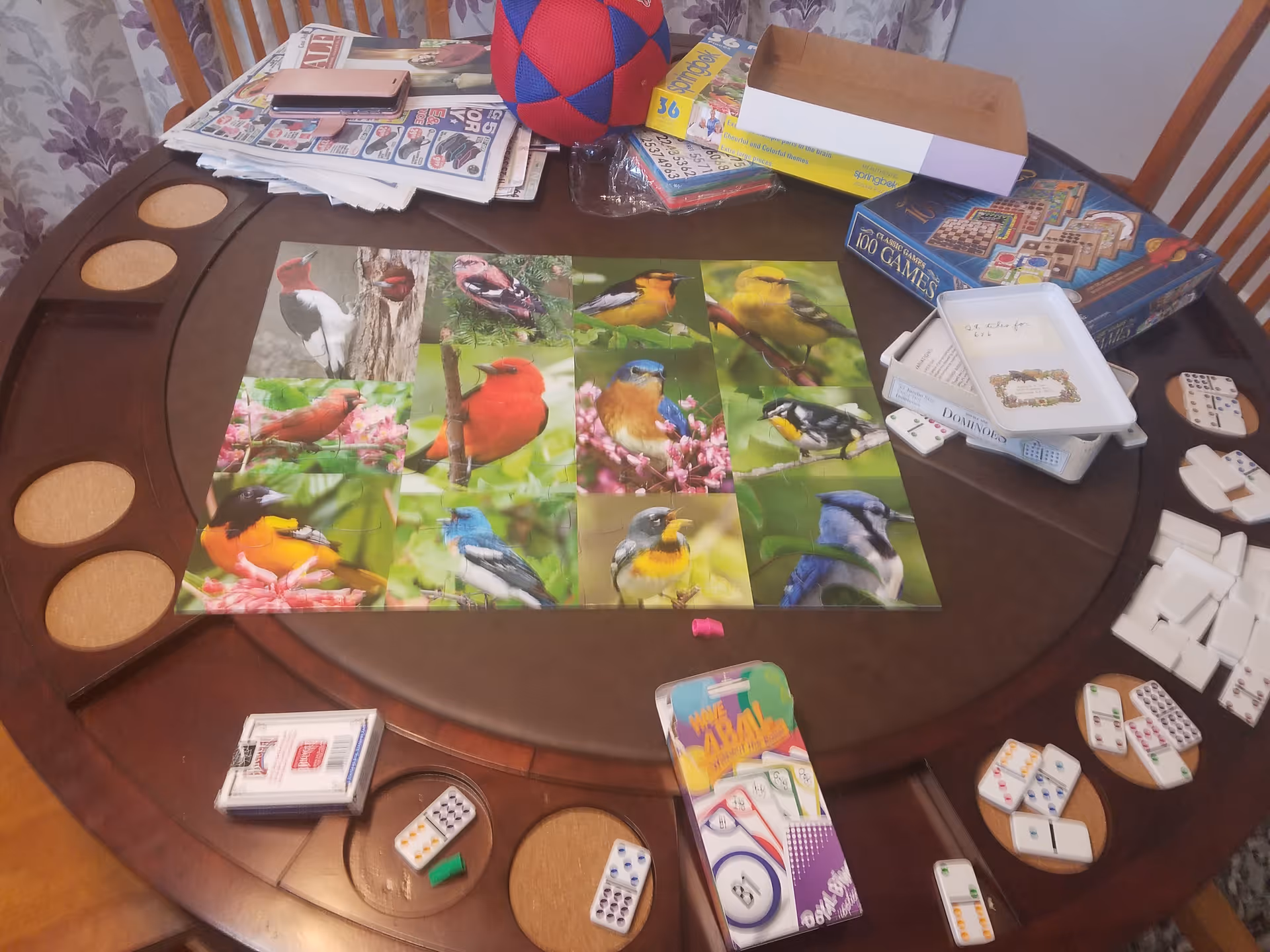 Round wooden table covered with bird picture cards, dominoes, playing cards, newspapers and game boxes in a dining area.