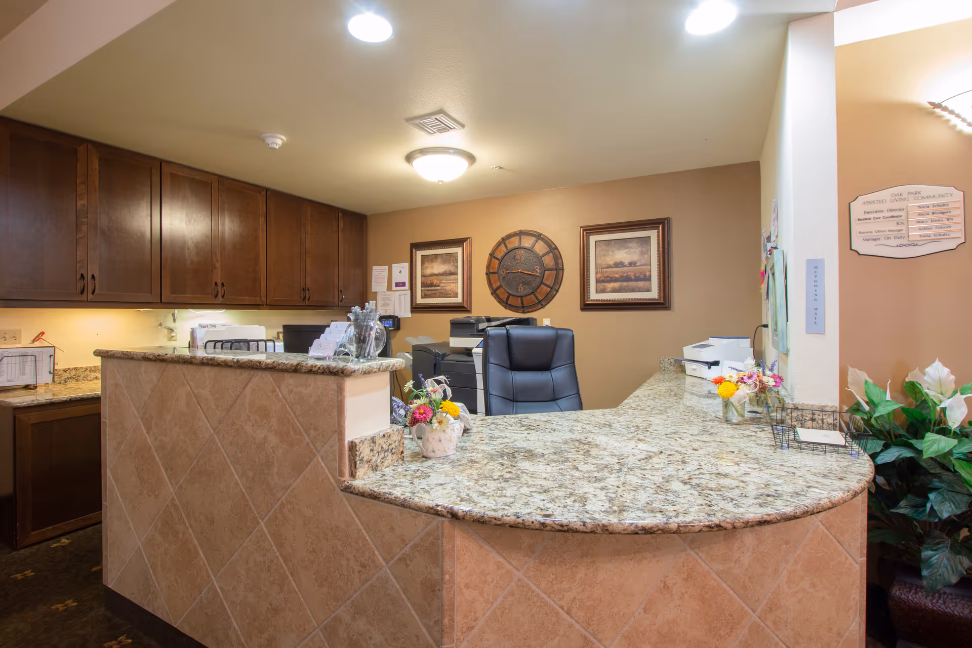 Reception desk area with a granite countertop and tiled front, a black office chair behind the desk, wooden cabinets on the wall, decorative flowers on the counter, a large wall clock, framed artwork, and a beige wall with a community information board.