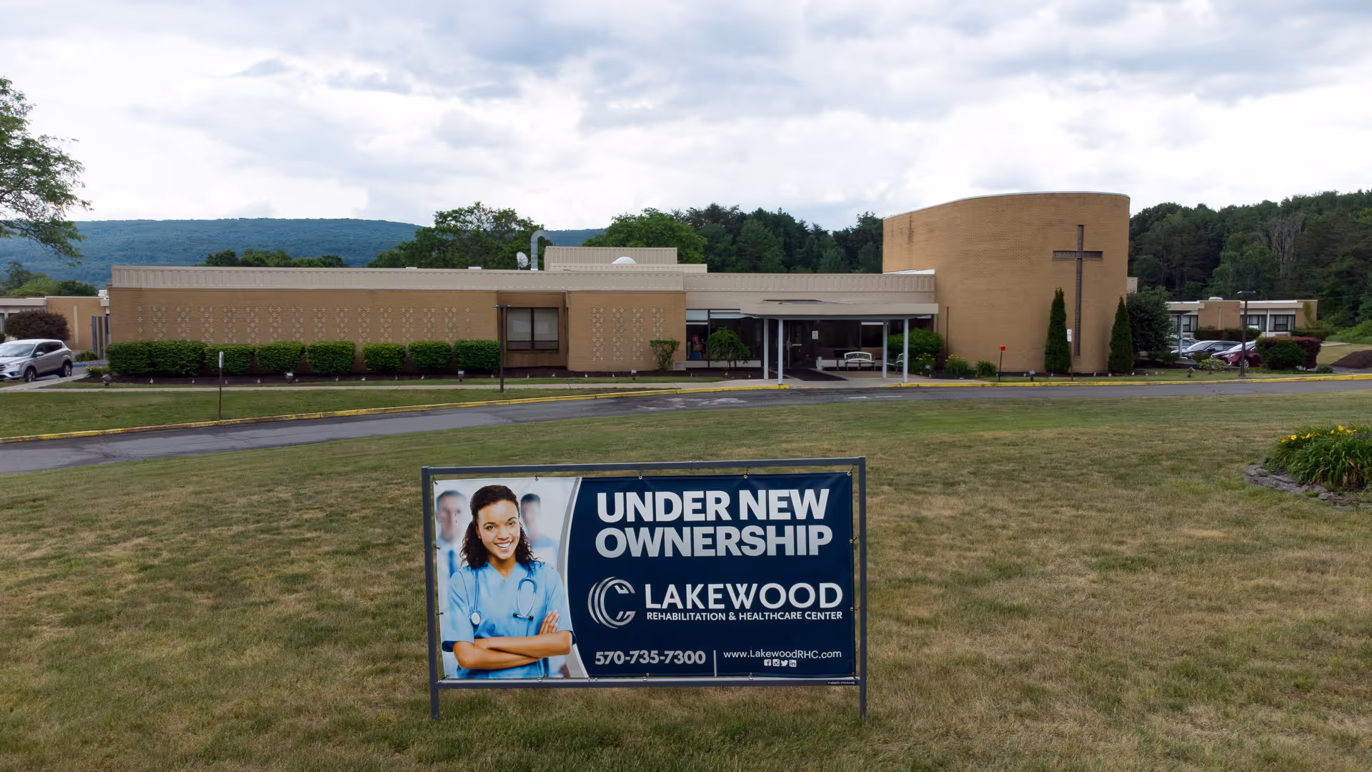 Exterior view of Lakewood Rehabilitation and Healthcare Center building with a large sign in the foreground that reads 'Under New Ownership' along with the facility's name, phone number, and website. The building is a single-story structure with a cylindrical section featuring a cross, surrounded by grass and trees with hills in the background under a cloudy sky.