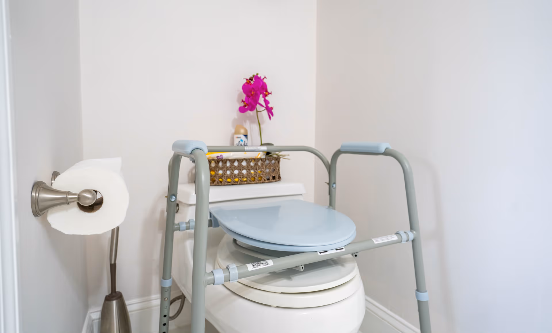 A bathroom scene showing a toilet with a raised toilet seat and safety frame around it. There is a toilet paper holder mounted on the wall to the left with a roll of toilet paper. On top of the toilet tank, there is a decorative basket and a purple orchid flower in a vase.
