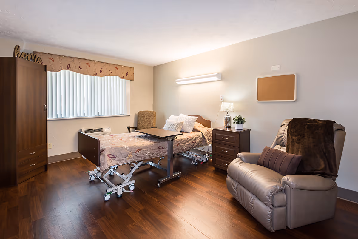 A senior living facility bedroom featuring a hospital-style adjustable bed with pillows and a patterned bedspread. Next to the bed is a wooden nightstand with a lamp and a small plant. There is a comfortable recliner chair with a brown throw blanket and pillow. The room has wooden flooring, a window with vertical blinds and a valance, a wooden wardrobe, and a cork bulletin board on the wall.