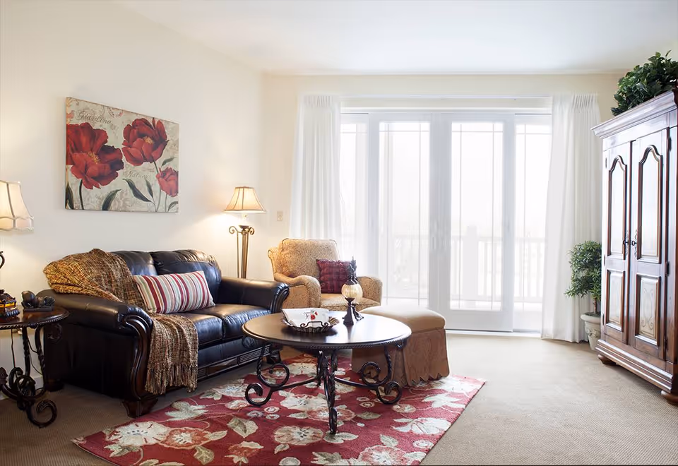 Bright living room with a leather sofa, armchair and ottoman, round coffee table on a floral rug, and large sliding glass doors.