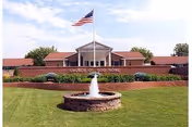 Front exterior view of the Church of God Home building with a flagpole flying the American flag, a circular stone fountain in the foreground, and well-maintained green lawn and shrubs.