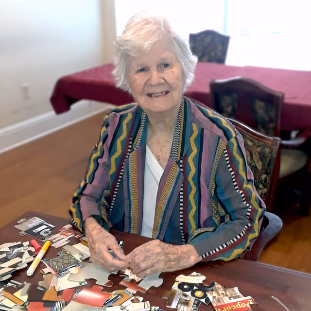An elderly woman with white hair wearing a colorful striped jacket is sitting at a wooden table working on a jigsaw puzzle. Behind her, there is a table covered with a red tablecloth and chairs in a well-lit room.