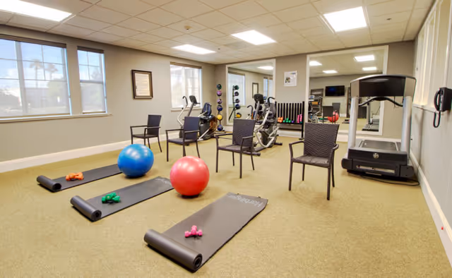 A fitness room with exercise equipment including a treadmill, stationary bikes, dumbbells, exercise balls, yoga mats, and chairs. The room has beige carpet, large windows, and mirrors on one wall.