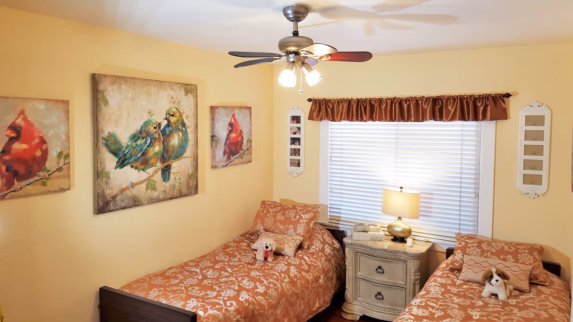 A cozy bedroom with two twin beds covered in matching orange floral bedspreads. Each bed has a stuffed animal placed on top. Between the beds is a vintage-style nightstand with a lamp, a book, and a small candle. The window behind the nightstand has white blinds and a brown valance. The walls are painted light yellow and decorated with three paintings of colorful birds and two white-framed photo displays. A ceiling fan with lights is mounted on the ceiling.