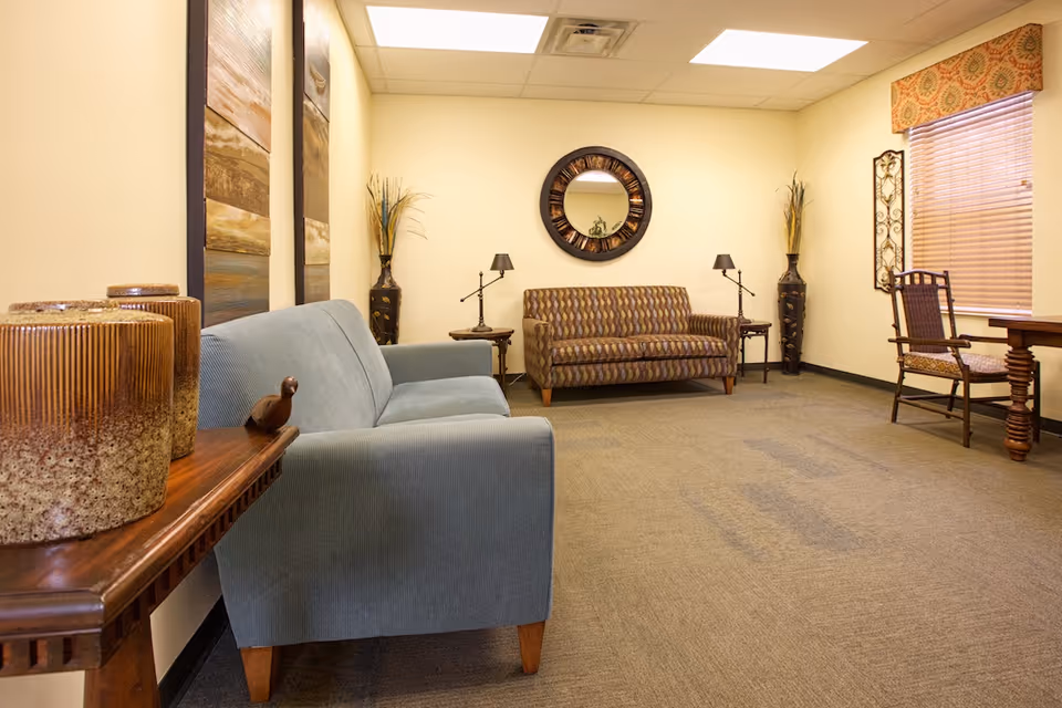 A cozy sitting area in a senior living facility with two sofas, a wooden side table with decorative vases, two small lamps on side tables, a round wall mirror, a window with blinds and a patterned valance, and a wooden chair next to a wooden table.