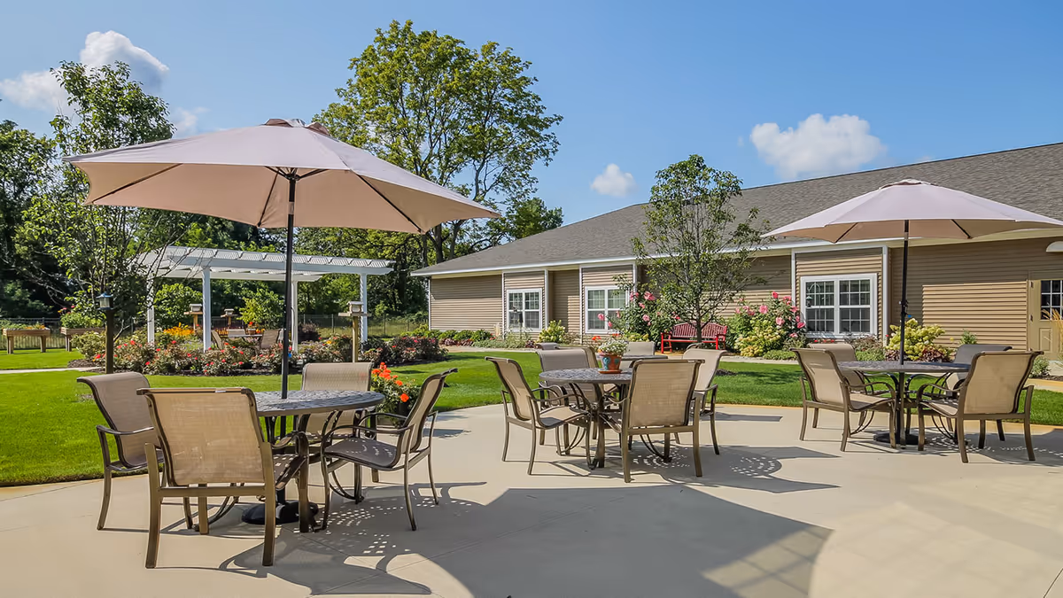 Outdoor patio area at Boulder Creek Assisted Living & Memory Care with round tables, chairs, and large beige umbrellas. The patio is surrounded by green grass, trees, and flowering plants with a building in the background under a clear blue sky.