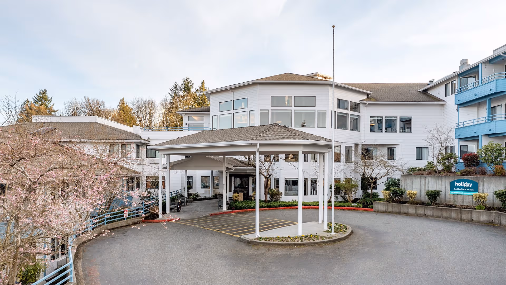 Exterior view of a multi-story senior living facility with white walls and blue balconies. The entrance has a covered driveway with a flagpole in the center. Trees and shrubs surround the building, and a sign near the entrance reads 'holiday CASCADIAN PLACE'.