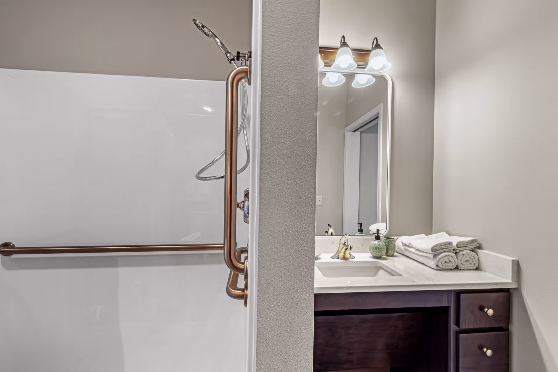 A bathroom with a white shower area featuring a metal grab bar and a handheld showerhead. Next to the shower is a vanity with a white countertop, a sink, a faucet, a soap dispenser, and neatly folded white towels. Above the vanity is a mirror with three light fixtures mounted on the wall.