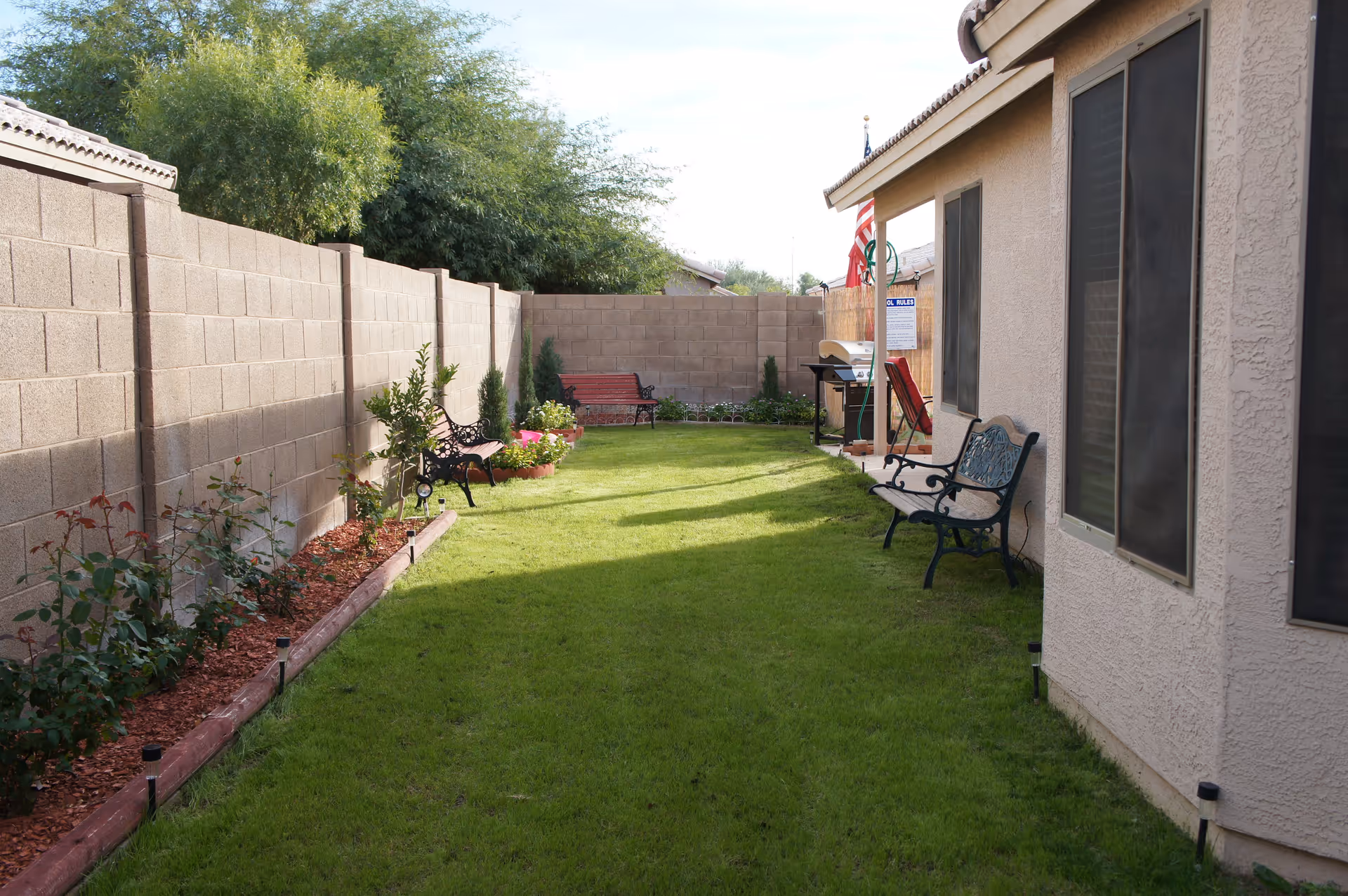 A narrow outdoor garden area with green grass, bordered by a beige concrete block wall on the left and a beige stucco building on the right. There are several benches along the sides, some plants and flowers along the wall, and an American flag near the building. A barbecue grill and a red chair are also visible near the building.
