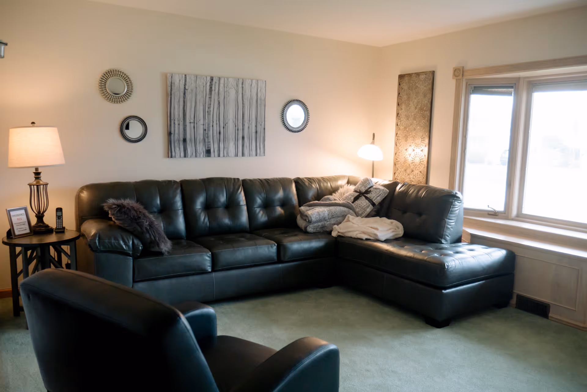 A cozy living room with a dark leather sectional sofa adorned with pillows and blankets, a matching armchair, a side table with a lamp and a cordless phone, three decorative round mirrors and a painting of birch trees on the wall, and a large window letting in natural light.