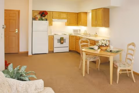 A small kitchen and dining area in a senior living facility. The kitchen has wooden cabinets, a white refrigerator, stove, microwave, and sink. A wooden dining table with two chairs is set with placemats, cups, and a basket of flowers. There is a beige carpet and a beige couch partially visible in the foreground.