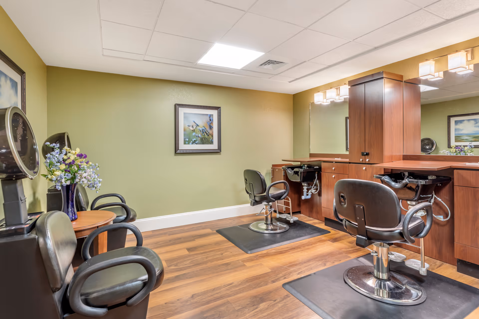 Interior of a salon area with two black salon chairs in front of washbasins and mirrors, wooden cabinetry, a small round table with a vase of flowers, and framed artwork on green walls.