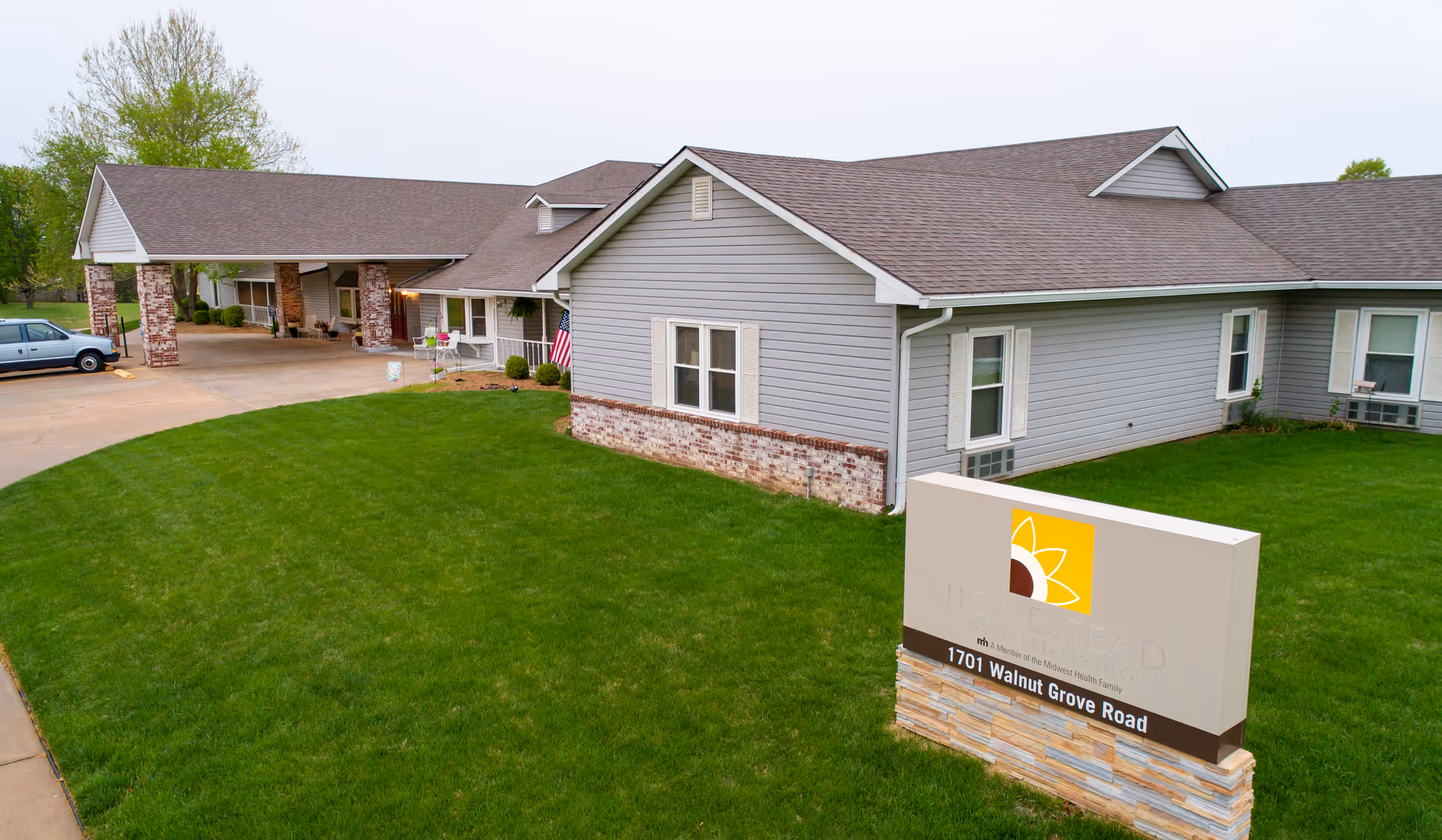 Exterior view of Homestead Assisted Living of Derby, showing a single-story building with gray siding and a brown shingled roof. There is a green lawn in front and a sign displaying the facility's name and address, 1701 Walnut Grove Road. A car is parked near the entrance, and an American flag is visible near the building.